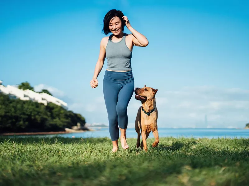Happy young woman and her pet dog running by the beach against clear blue sky, playing and enjoying time together in the nature outdoors. Living with a dog. Love and bonding with pet